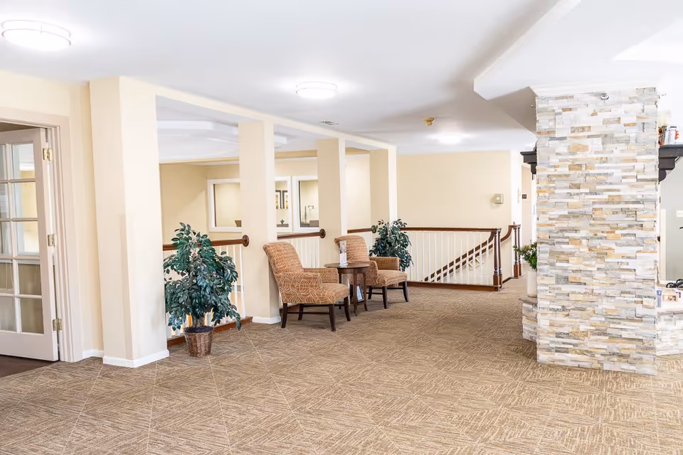 A bright and spacious interior hallway area with beige walls and carpeted floor. Two patterned armchairs with a small round table between them are placed near a railing overlooking a staircase. There are potted plants on either side of the chairs and a stone pillar on the right side of the image. The area is well-lit with ceiling lights and has a calm, welcoming atmosphere.
