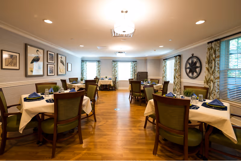 Bright communal dining room with round tables set with place settings, green upholstered chairs, wood floors, and decorative wall art.