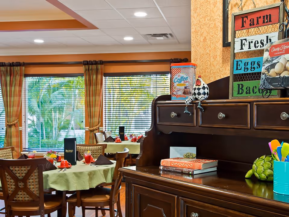 Interior view of a dining room in a senior living facility with round tables covered in light green tablecloths, set with red cups, napkins, and menus. Large windows with plaid curtains allow natural light to brighten the room. In the foreground, a dark wooden sideboard holds decorative items including a rooster figurine, cookbooks, and a colorful sign that reads 'Farm Fresh Eggs Bacon'.