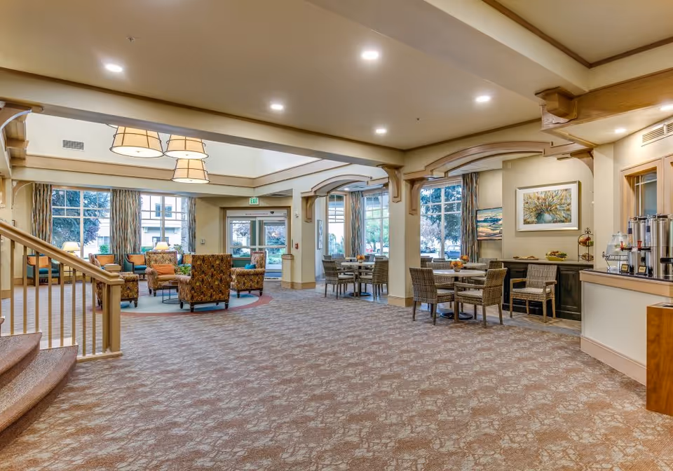 A spacious and well-lit common area in a senior living facility featuring a seating area with armchairs and a coffee table on the left, several dining tables with wicker chairs on the right, large windows with curtains allowing natural light, and a beverage station with coffee dispensers on the far right.