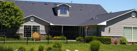 Exterior view of a single-story residential building with beige siding, a dark shingled roof, and multiple windows. The building is surrounded by a well-maintained lawn, shrubs, and a black metal fence. There is a wooden bench on the patio area near the building.