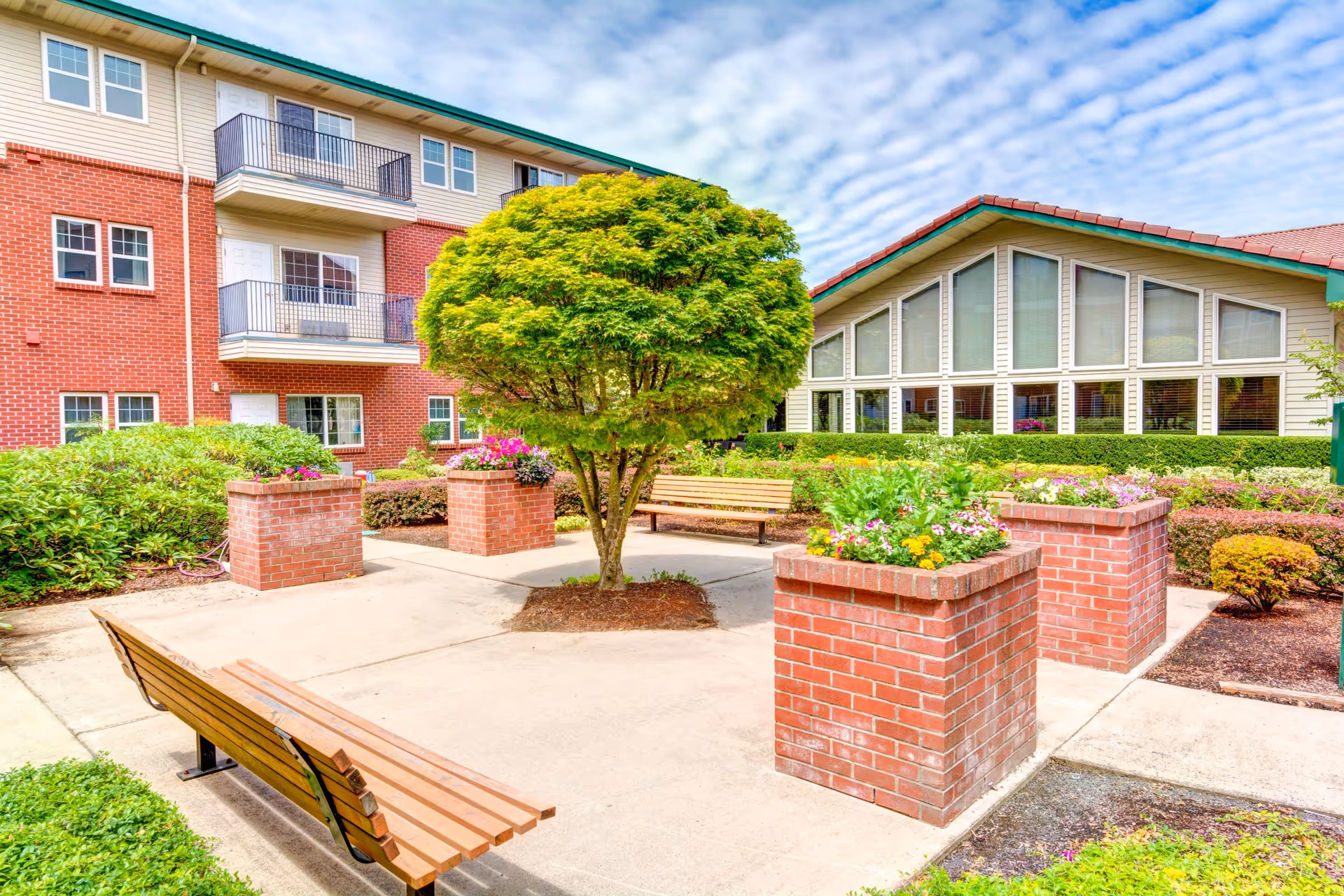 Outdoor courtyard area at Rosewood Park featuring a central tree surrounded by brick planters with colorful flowers, wooden benches, and well-maintained shrubs. The background shows a multi-story brick and siding building with balconies and a large building with many windows under a partly cloudy sky.