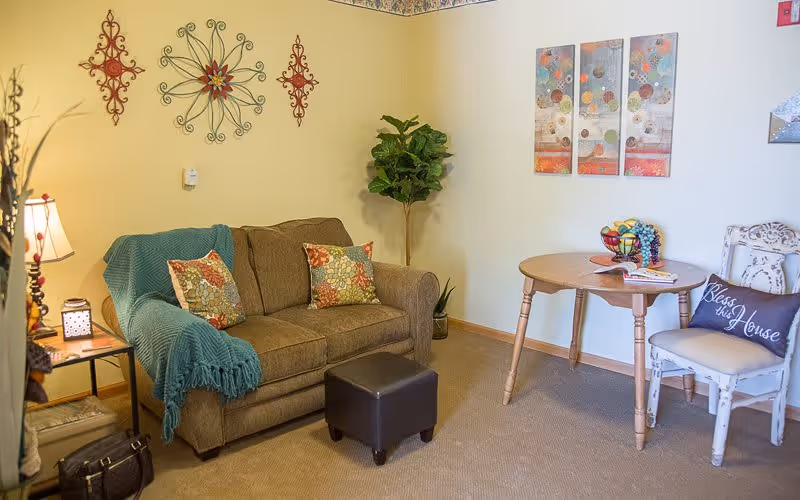 A cozy living room area with a brown sofa adorned with colorful floral pillows and a teal throw blanket. A small brown ottoman is placed in front of the sofa. To the right, there is a round wooden table with a fruit basket and some magazines on it, accompanied by a white distressed chair with a pillow that reads 'Bless this House'. The walls are decorated with metal wall art and a three-panel abstract painting. A potted plant is in the corner, and a side table with a lamp and decorative items is next to the sofa.