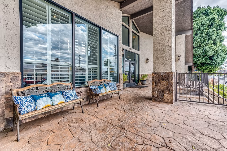 Outdoor covered patio area at the entrance of a building with two wooden benches adorned with blue and white floral cushions, large windows with white shutters, stone and stucco walls, and a metal gate on the right side.