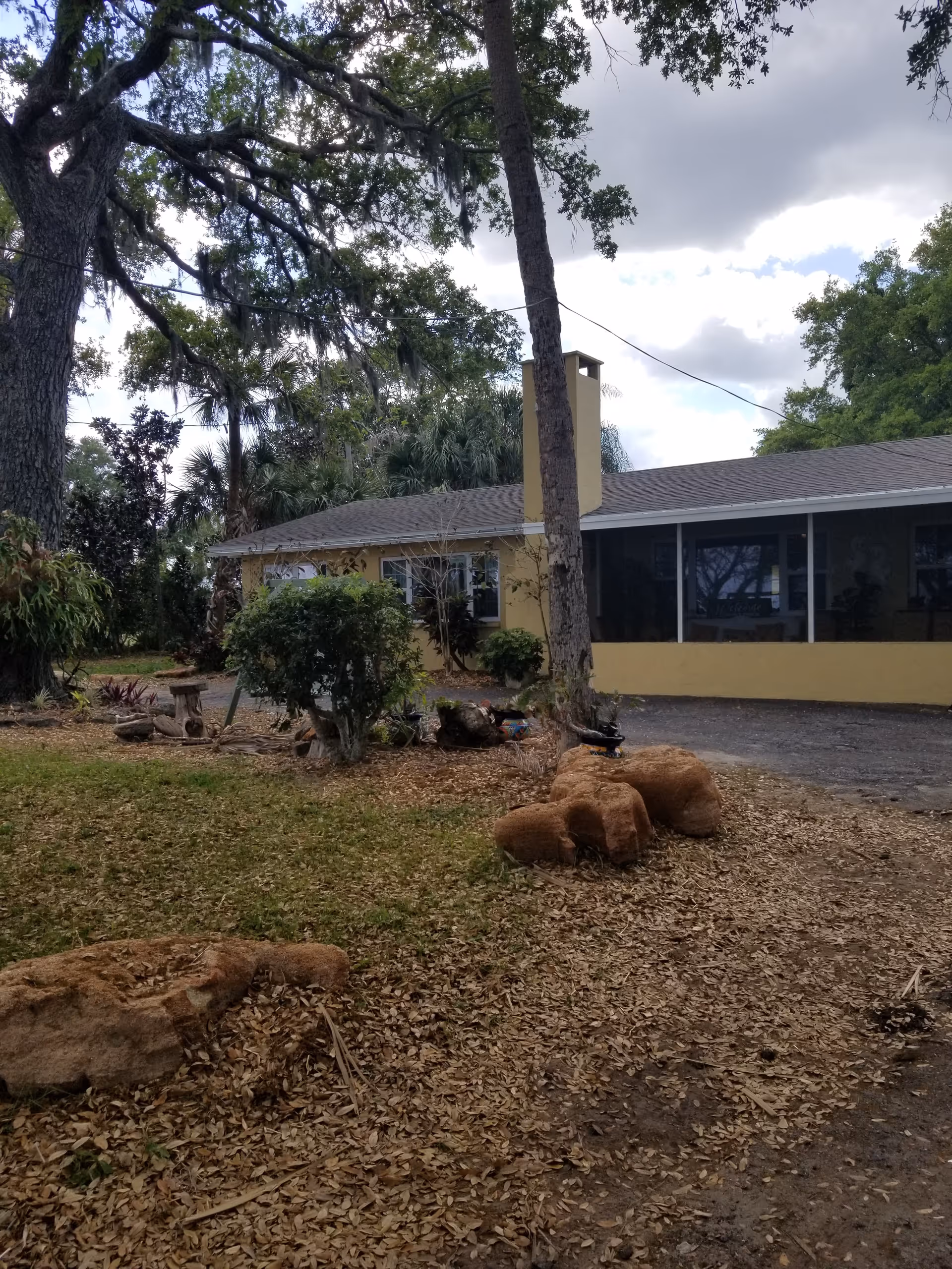 Exterior view of a single-story yellow building with a screened porch, surrounded by trees and bushes under a cloudy sky. The ground is covered with dry leaves and some large rocks are visible in the foreground.