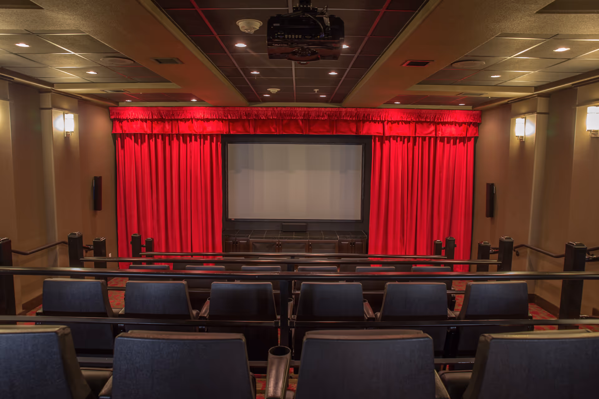 Interior view of a small theater room with rows of blue cushioned seats facing a large screen framed by red curtains. The room has beige walls with wall-mounted lights and a ceiling-mounted projector.