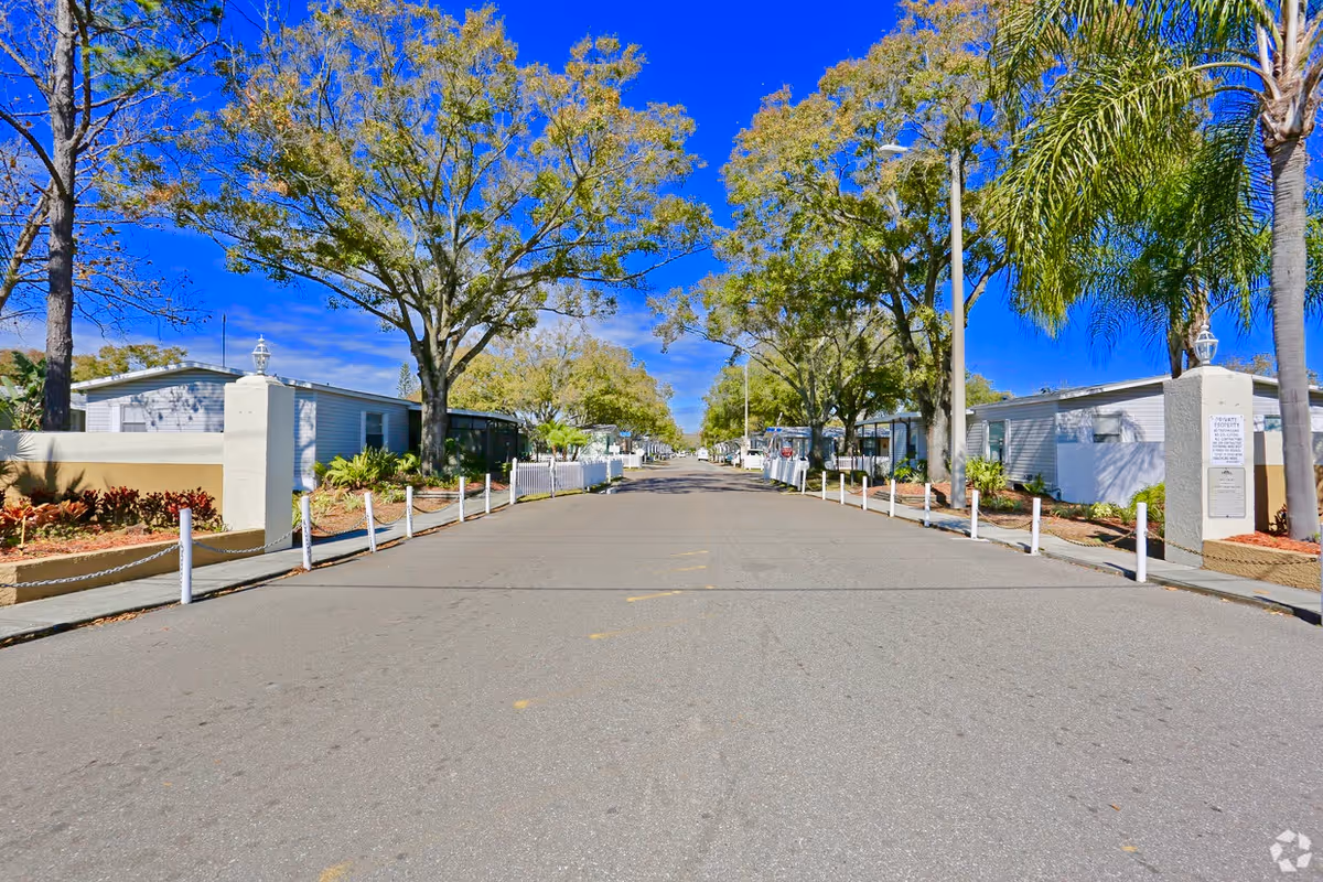 A wide paved road lined with white posts and chains on both sides, flanked by single-story residential buildings and trees under a clear blue sky.
