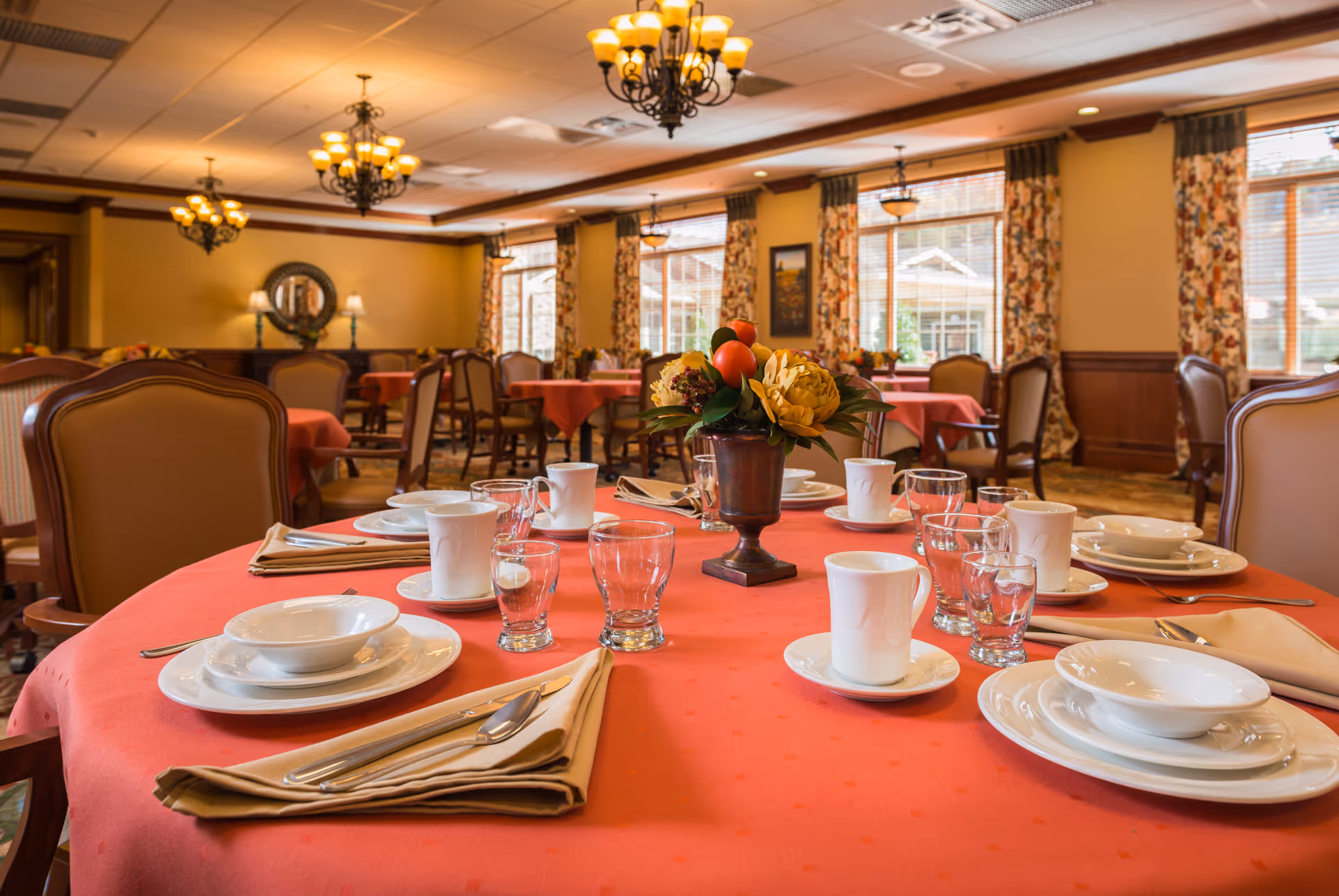 A dining room with round tables covered in red tablecloths, set with white plates, bowls, cups, glasses, and beige napkins. Each table has a floral centerpiece. The room features large windows with floral curtains, wooden paneling, and chandeliers hanging from the ceiling.