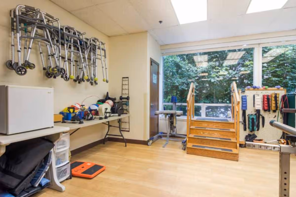 A rehabilitation or therapy room with multiple walkers hanging on the wall, a table with various therapy and exercise equipment, a small set of wooden stairs for physical therapy, and large windows showing greenery outside.