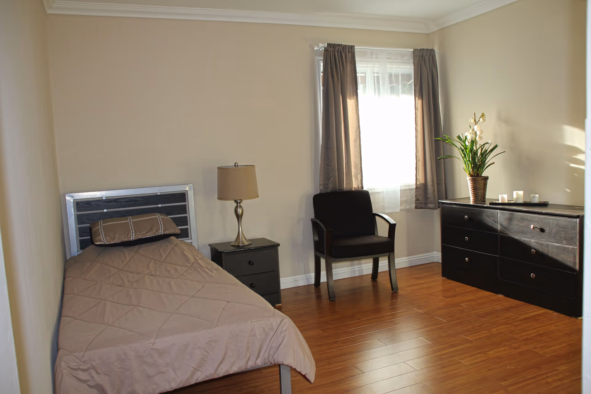 A simple bedroom with a single bed covered in a beige quilt, a matching pillow, a small black nightstand with a lamp, a black armchair, a window with gray curtains, and a black dresser with a potted plant and candles on top. The room has wooden flooring and beige walls.