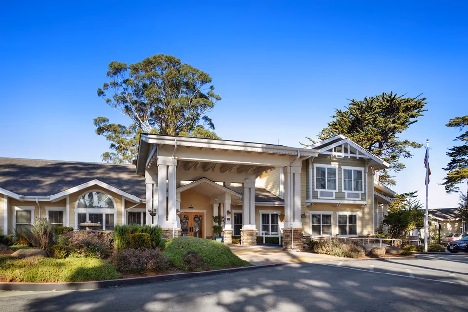 Front exterior of a senior living facility with a covered entrance, landscaped beds, and large windows under a clear blue sky.