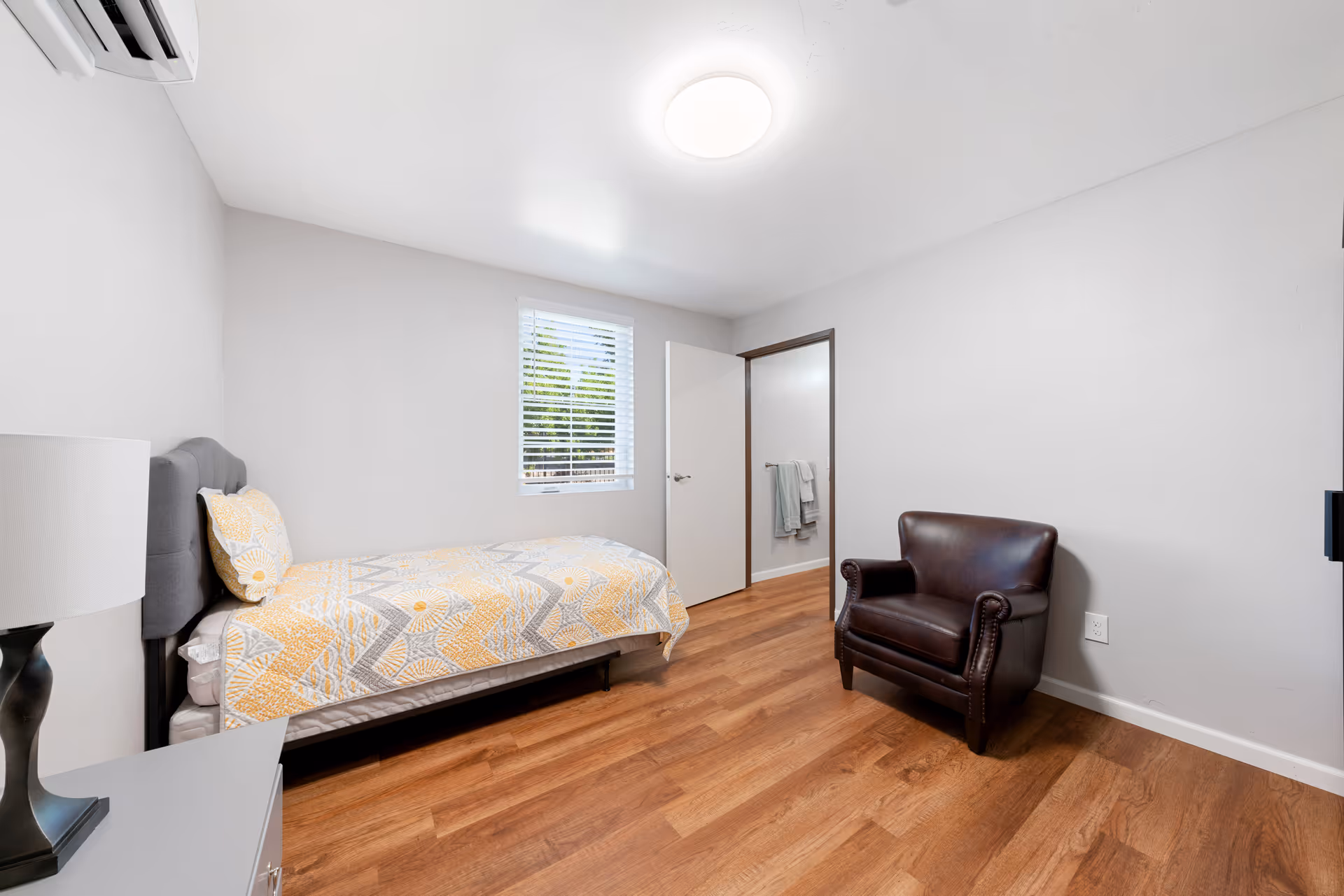 Small furnished bedroom with a single bed, bedside lamp, leather armchair, window with blinds, and an open door.