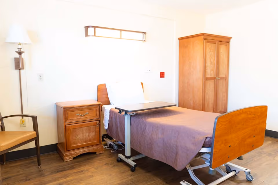 A simple, clean room with a single hospital-style bed covered with a brown blanket, a white pillow, and an adjustable overbed table. Next to the bed is a wooden nightstand with a drawer and cabinet, a wooden wardrobe, a wooden chair with a tan cushion, and a wall-mounted lamp. The floor is wooden, and the walls are plain white.
