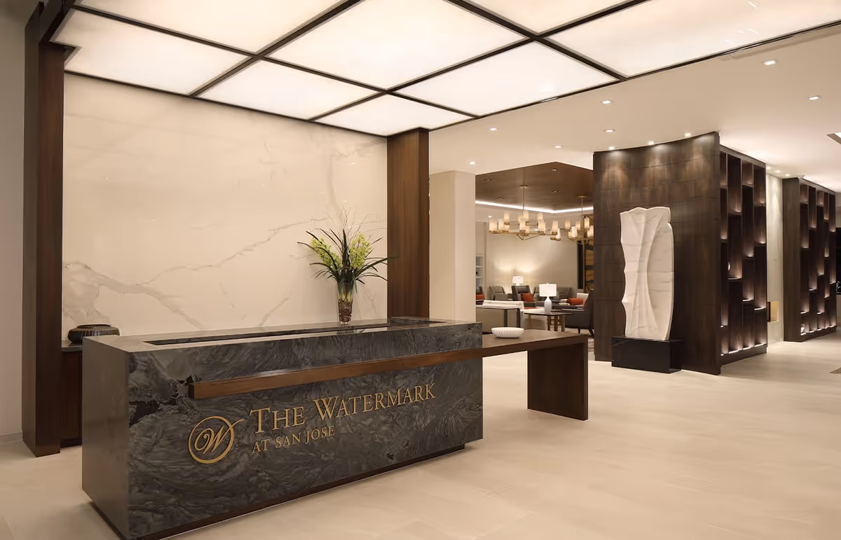 A modern and elegant reception area with a marble front desk displaying the text 'The Watermark at San Jose'. The space features a large lighted ceiling, a decorative vase with flowers on the desk, a sculptural art piece on a pedestal, and a seating area with chairs and lamps in the background.