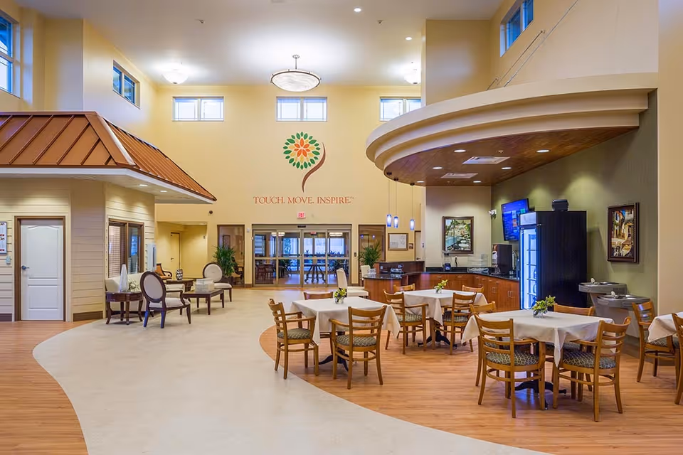 Interior view of a senior living facility common area with tables and chairs arranged for dining. The room features a high ceiling with multiple windows, a small seating area with armchairs, a counter with a beverage station, and decorative wall art including a colorful flower logo and the words 'TOUCH. MOVE. INSPIRE.'