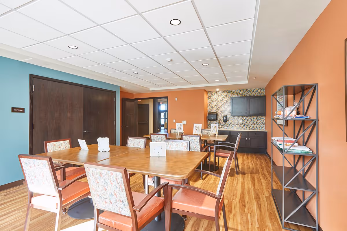 A bright and clean dining room with wooden tables and chairs featuring floral upholstery. The room has orange and teal walls, a wooden floor, and a ceiling with recessed lighting. There is a shelving unit on the right side with books and decorative items, and a kitchenette area with dark cabinets and a mosaic tile backsplash in the background.