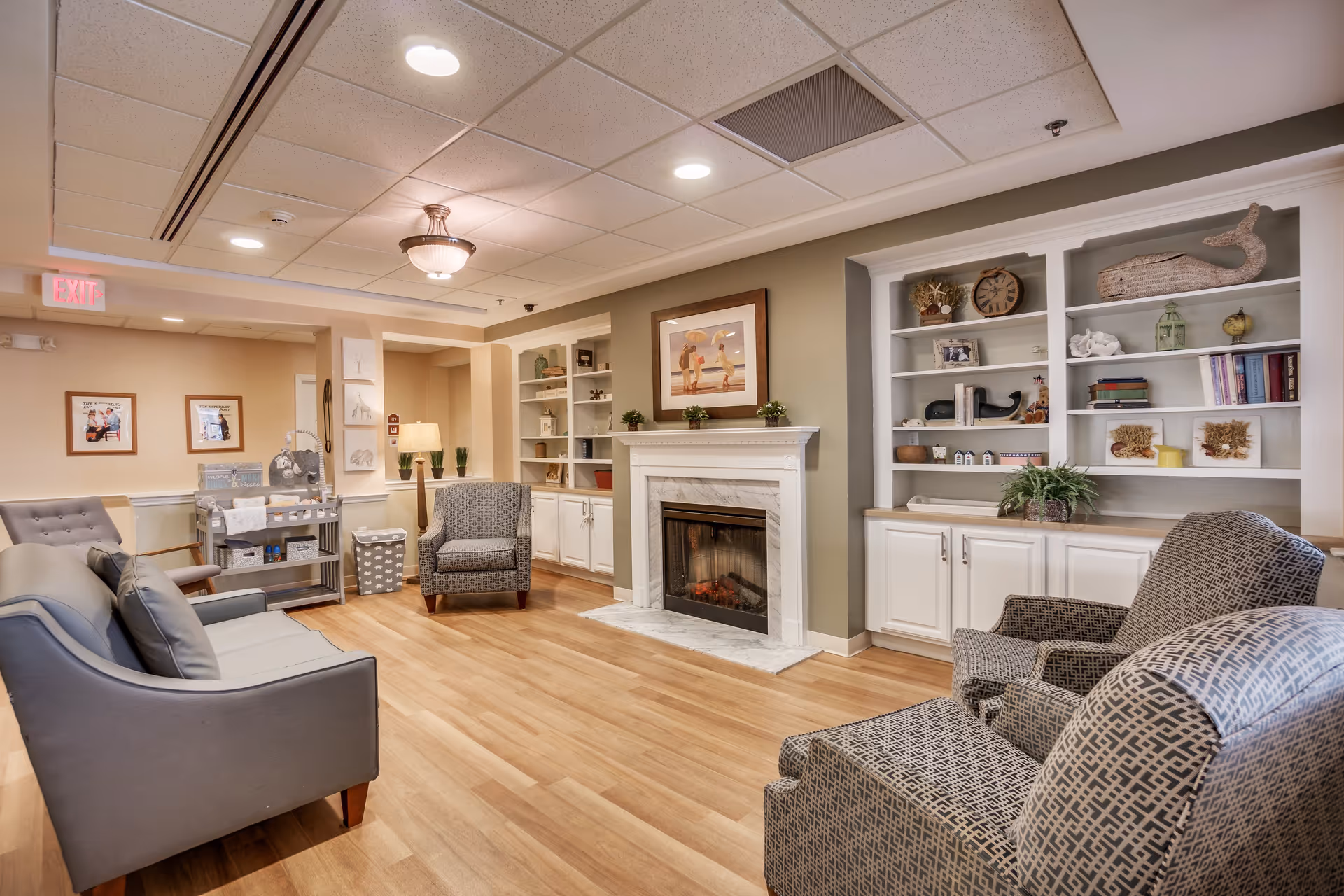 A cozy living room area in an assisted living facility featuring a fireplace with a framed picture above it, built-in white shelves with decorative items, two patterned armchairs, a gray sofa, wooden flooring, and soft lighting from ceiling fixtures and a floor lamp.