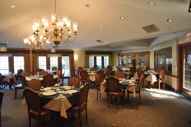 Dining room with multiple round tables set with plates and cups, upholstered chairs, chandeliers, and large windows.