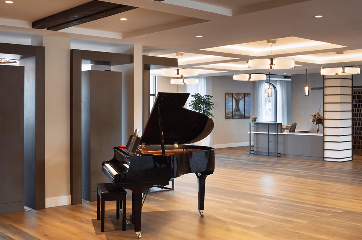 Spacious senior living facility common area with a black grand piano and matching bench on a wooden floor. The room features modern ceiling lights, a reception desk in the background, decorative wall art, and large windows with curtains allowing natural light.