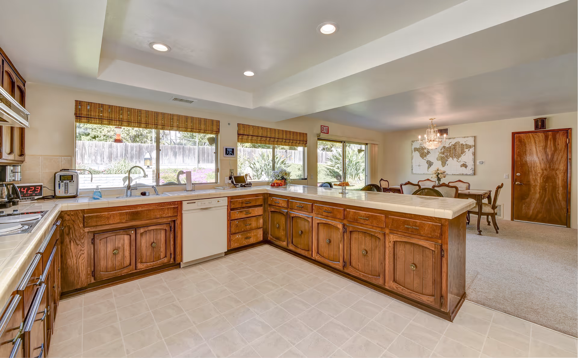 A spacious kitchen with wooden cabinets and a large countertop island. The kitchen has a dishwasher, a sink under large windows with bamboo blinds, and various kitchen appliances on the counters. Beyond the kitchen is a dining area with a wooden dining table, chairs, a chandelier, and a world map on the wall. There is a door and sliding glass doors leading outside.