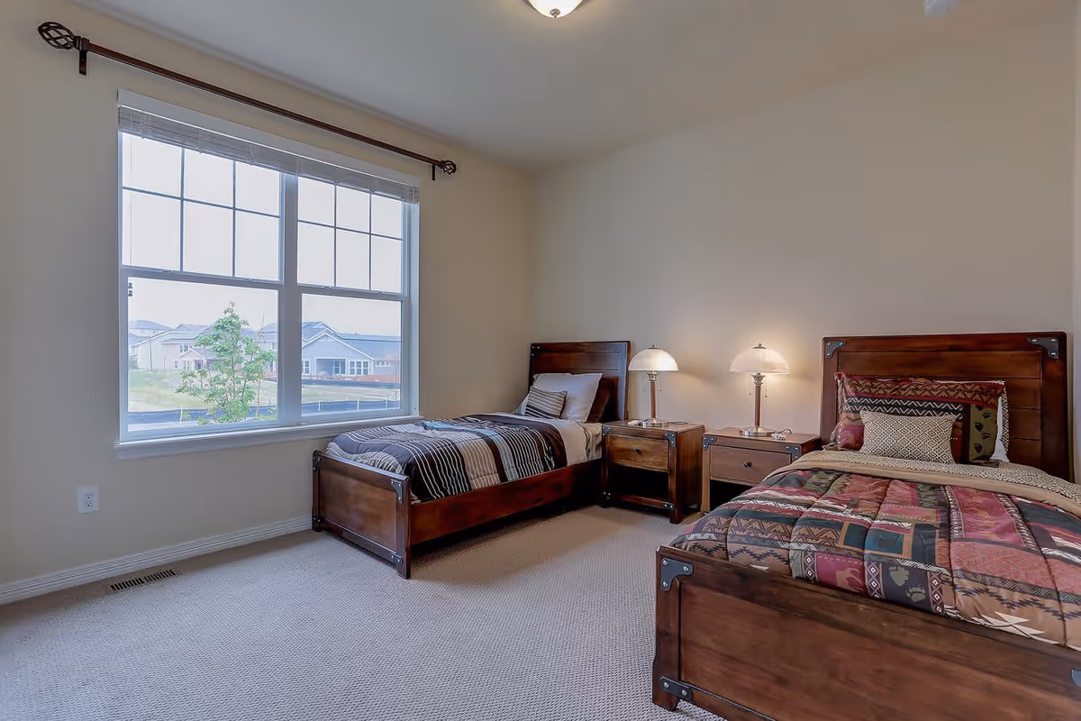 A bright bedroom with two single wooden beds, each with patterned bedding and pillows. Between the beds is a wooden nightstand with two lamps. A large window on the left side lets in natural light and shows a view of houses and greenery outside.