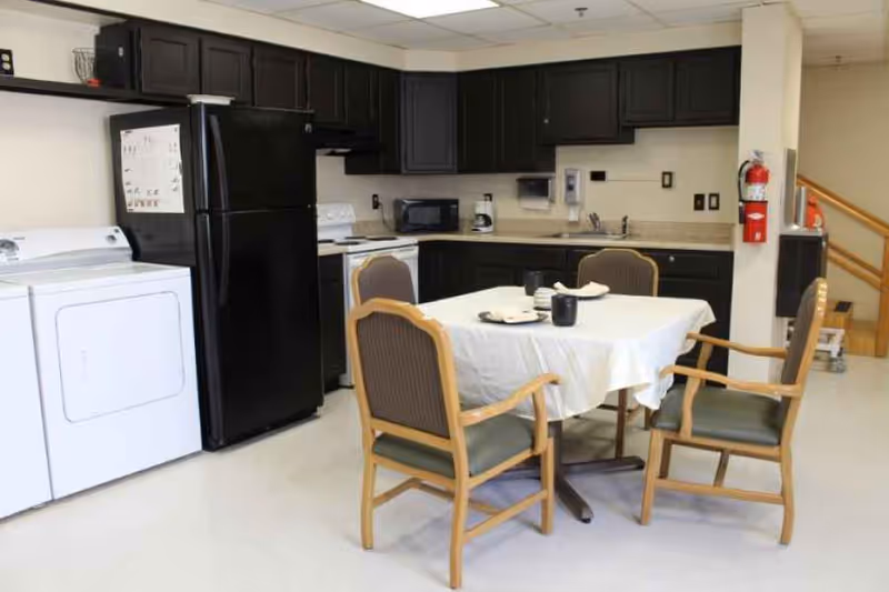 Small communal kitchen and dining area with a table and four chairs, black appliances, dark cabinets, and a washer and dryer.