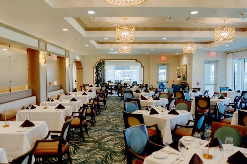 Spacious, elegantly set dining room with multiple tables covered in white tablecloths and hanging chandeliers.