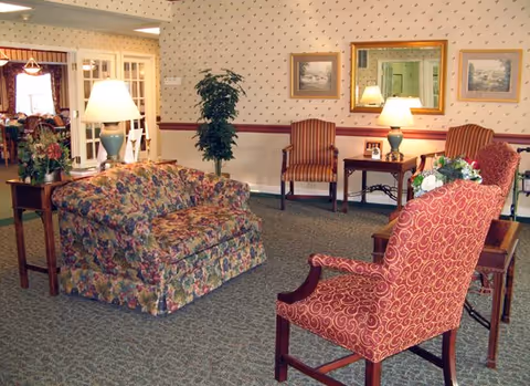 A carpeted common room with patterned armchairs, a floral loveseat, side tables with lamps, and framed artwork on the wall.