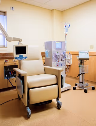 A medical treatment room with a beige reclining chair on wheels, a dialysis machine with attached IV bag, and a medical monitor on a stand. The room has light-colored walls and a window with blinds.
