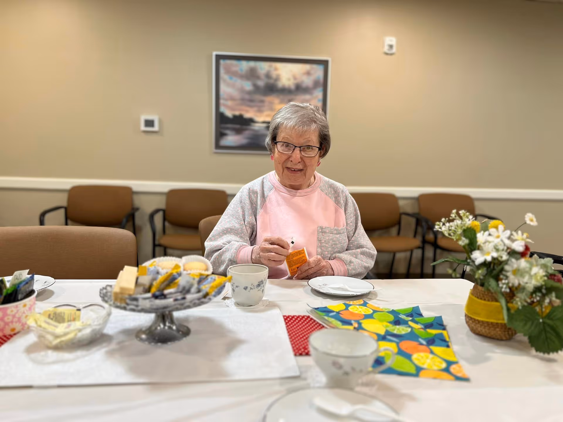 An elderly woman with glasses sitting at a table in a room with beige walls and brown chairs. The table is set with cups, plates, a bowl of sugar packets, a tiered tray with snacks, colorful napkins, and a small flower arrangement. A framed picture hangs on the wall behind her.