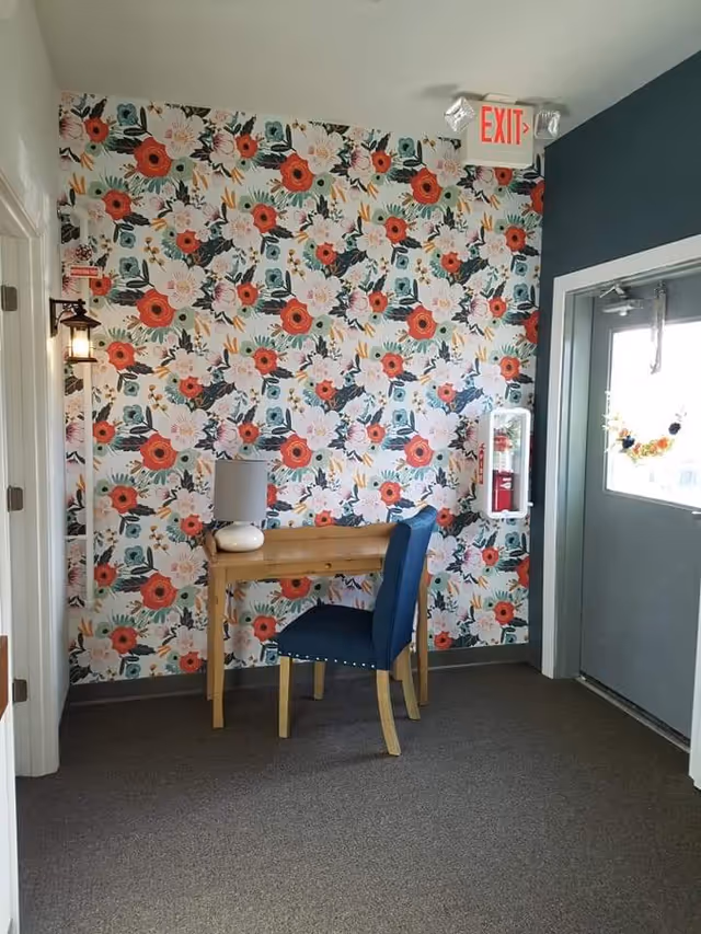 A small corner area inside a facility with a floral patterned wallpaper featuring large red, white, and pink flowers. There is a wooden desk with a white lamp on it and a blue upholstered chair in front of the desk. To the right, there is a gray door with a window decorated with a small floral wreath. Above the door is an illuminated red EXIT sign. A fire extinguisher is mounted on the wall next to the door.