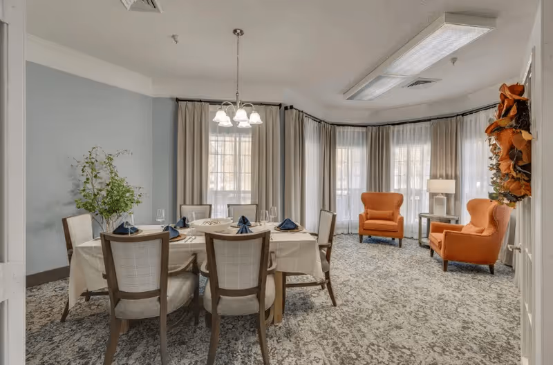 A dining room with a rectangular table covered with a white tablecloth and set with blue napkins and glassware. Six chairs surround the table. In the background, there are large windows with sheer curtains and beige drapes. Two orange armchairs and a side table with a lamp are positioned near the windows. A decorative wreath hangs on the right wall.