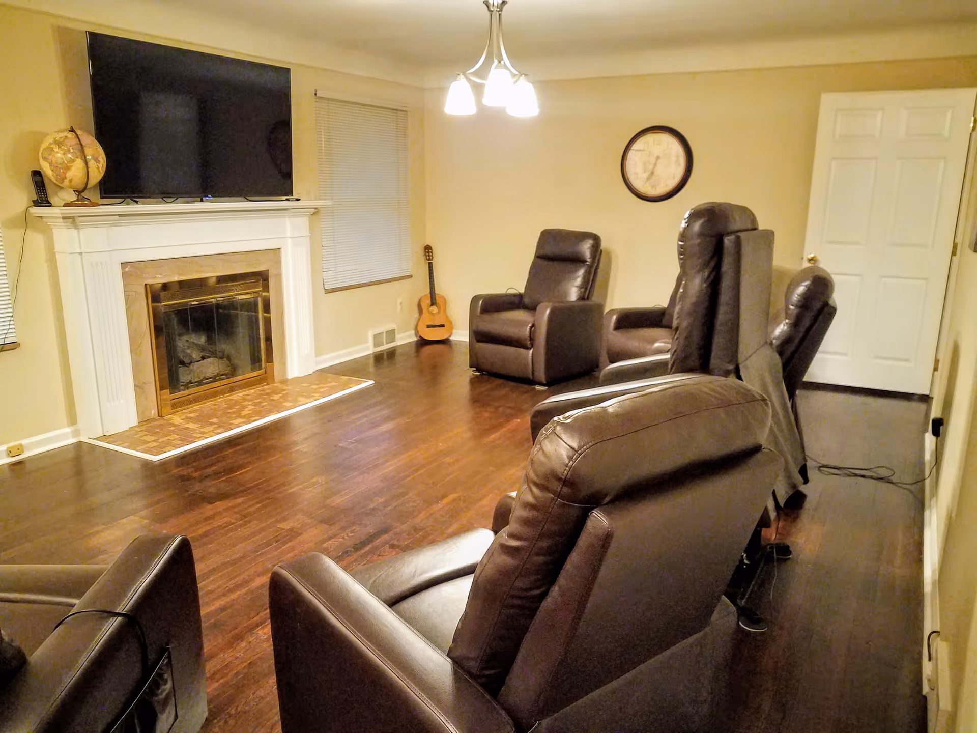 A cozy living room with four brown leather recliners arranged facing a white fireplace with a mounted flat-screen TV above it. There is a globe and a cordless phone on the fireplace mantel. A guitar is placed in the corner near a window with closed blinds. A round wall clock hangs on the beige wall, and a white door is visible in the background. The room has wooden flooring and a ceiling light fixture with three bulbs.
