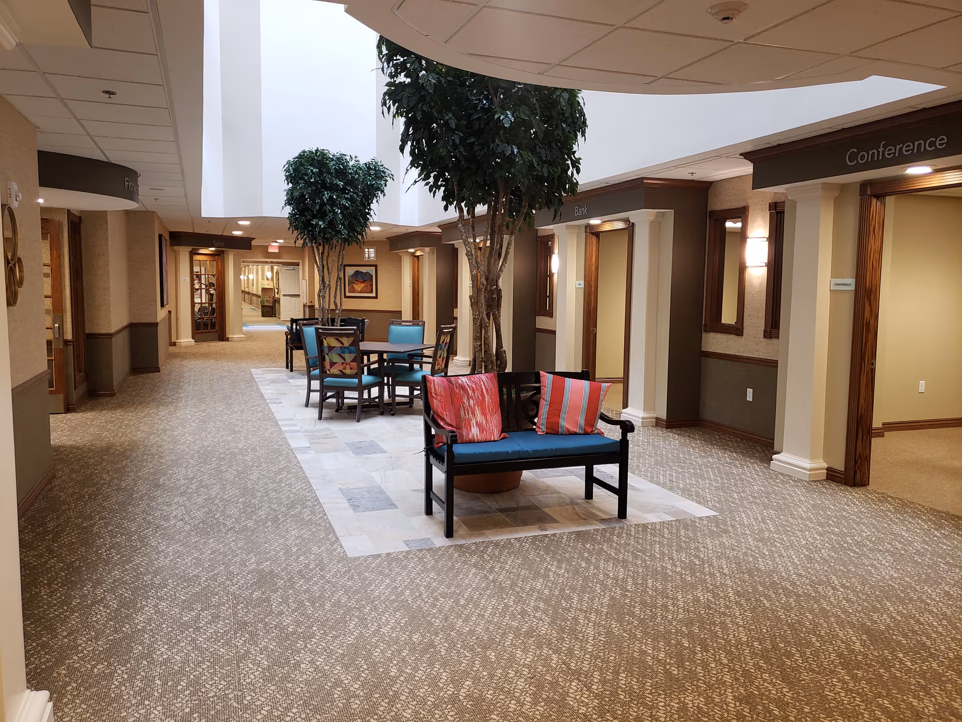 Interior hallway of a retirement community with carpeted floors and a tiled seating area in the center featuring a bench with colorful cushions and a table with chairs. There are tall indoor plants and doorways labeled 'Bank' and 'Conference'. The ceiling has a skylight allowing natural light to brighten the space.