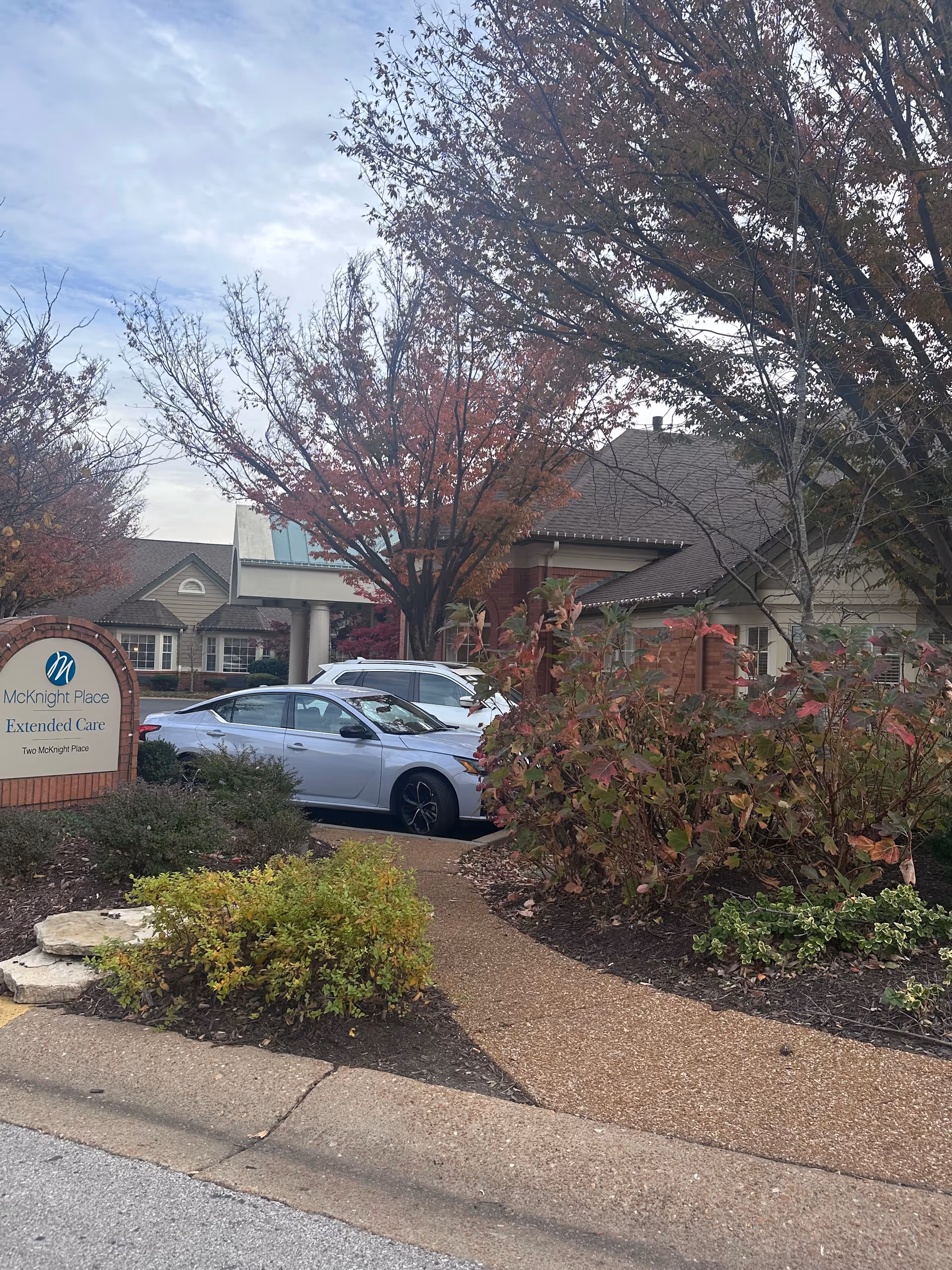 Exterior view of McKnight Place Extended Care facility showing a brick sign with the facility name, a pathway surrounded by bushes and trees with autumn foliage, and parked cars near the building under a cloudy sky.