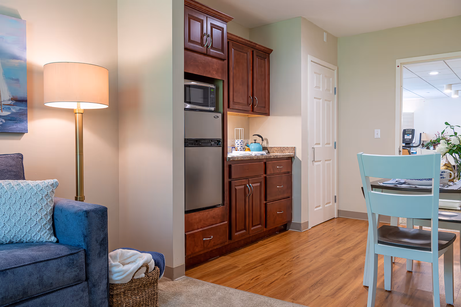 Interior view of a senior living facility showing a cozy living area with a blue sofa and a floor lamp, adjacent to a kitchenette with wooden cabinets, a microwave, and a small refrigerator. A light blue chair and part of a dining table are visible on the right side, with a doorway leading to another room in the background.