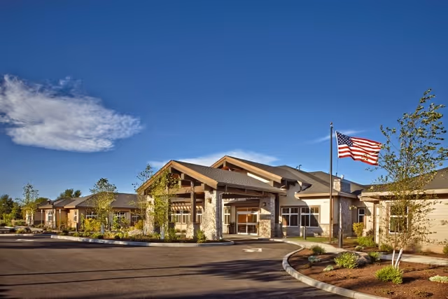 Front exterior of a single-story senior care building with a covered porte-cochère, landscaped grounds, and an American flag under a clear blue sky.