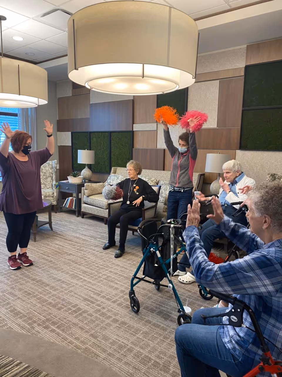 A group of elderly individuals and a caregiver participating in a seated exercise or activity session in a well-lit common room. One caregiver stands with arms raised, while another person holds orange and pink pom-poms. The room has modern decor with large hanging lights, comfortable chairs, and a carpeted floor.