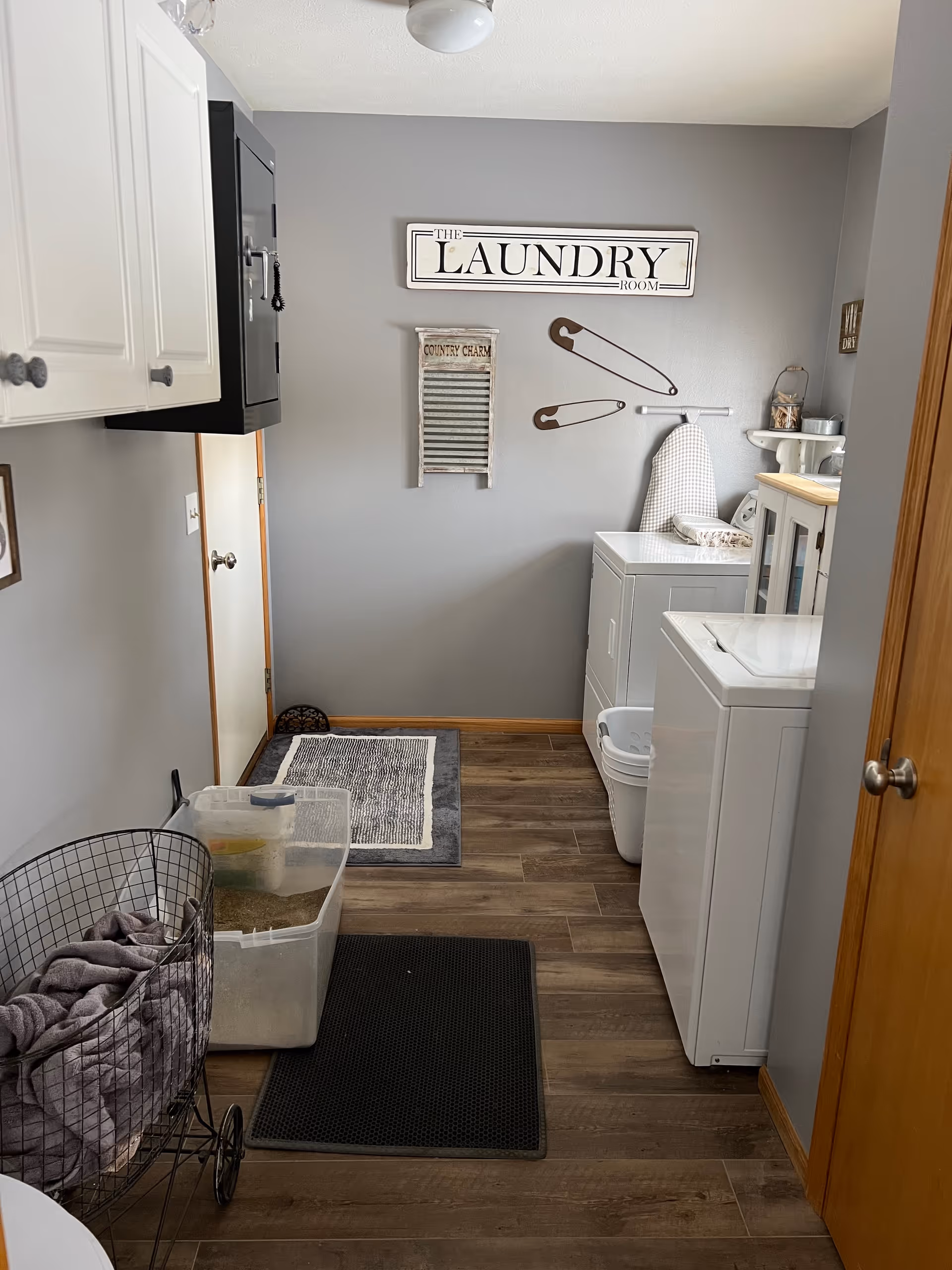 Laundry room with a washing machine and dryer on the right side, a wire laundry basket with clothes on the left, and decorative wall signs including a large sign that says 'The Laundry Room' and oversized safety pins. The room has gray walls, wood flooring, and a wooden door on the right.