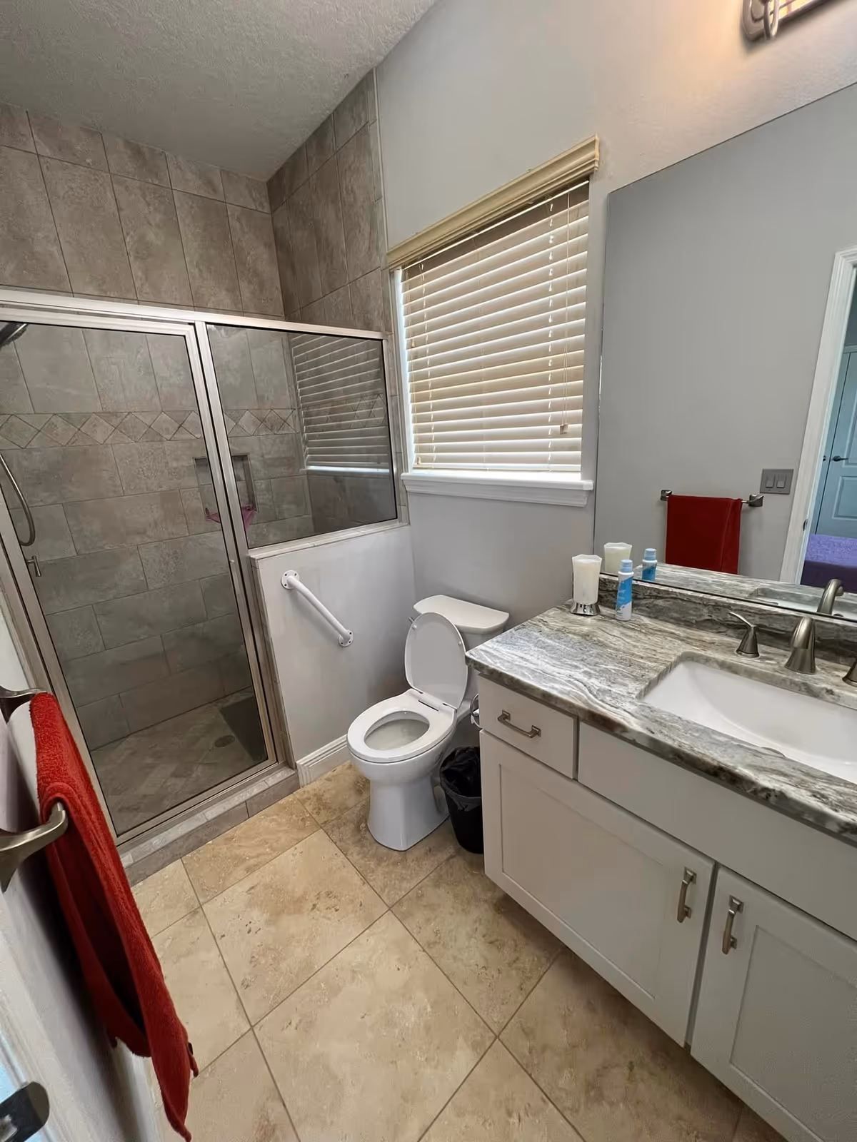 A bathroom with a tiled walk-in shower with glass doors, a white toilet with the lid open, a vanity with a marble countertop and an undermount sink, a large mirror above the vanity, a window with closed blinds, and red towels hanging on a towel rack.
