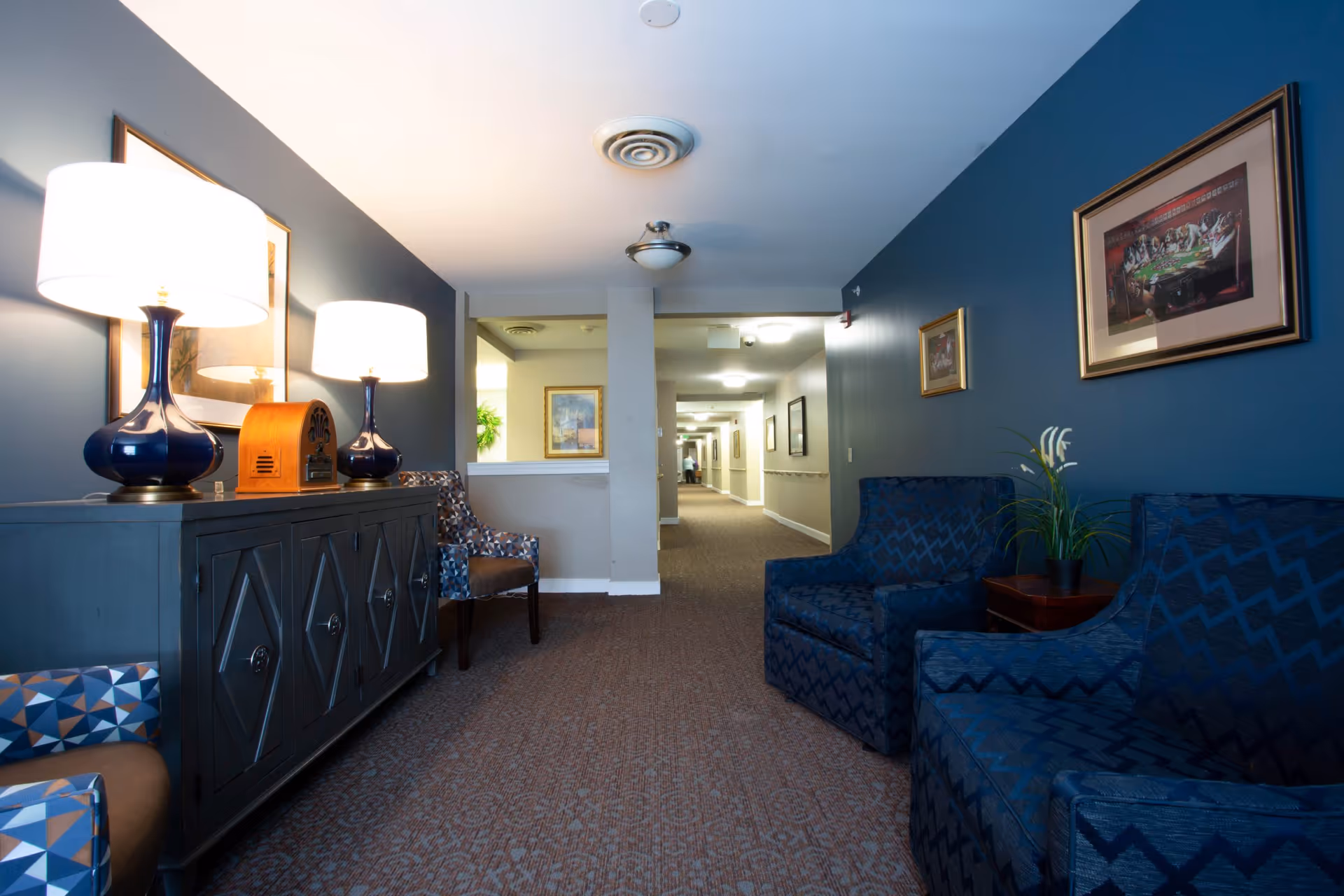 Seated lounge area in a senior living facility hallway with blue armchairs, a sideboard topped with lamps, and framed artwork.