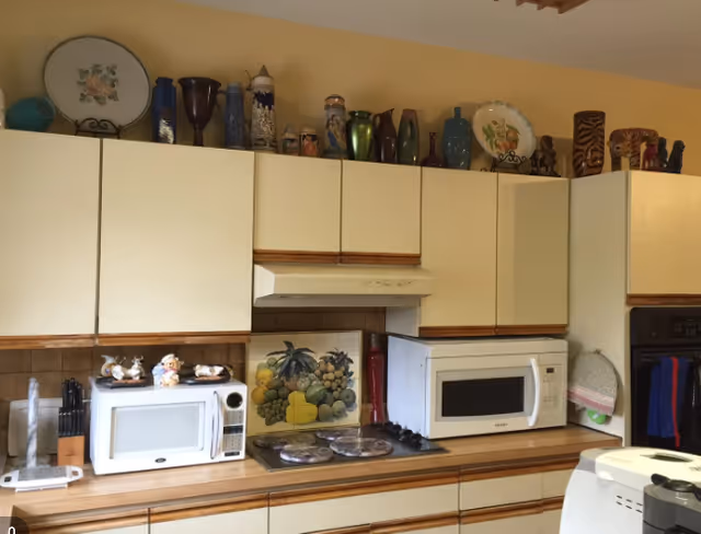 A kitchen interior with white cabinets and wooden trim. The countertop holds two microwaves, a stovetop with four burners, and various kitchen utensils. Decorative plates and vases are displayed on top of the cabinets. The wall behind the stovetop features a tile mural with fruit designs.