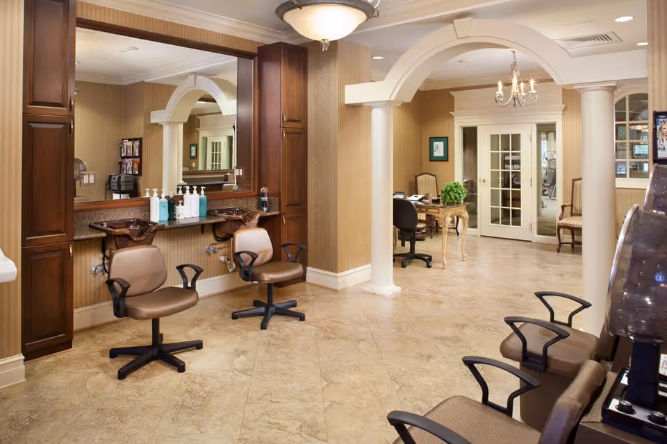 Interior view of a salon area in The Woodlands Retirement Community featuring two brown salon chairs in front of a large mirror with hair washing sinks. The room has beige tiled flooring, beige walls, and white decorative columns. In the background, there is a desk with a chair and a green plant, and a set of glass double doors leading to another room.