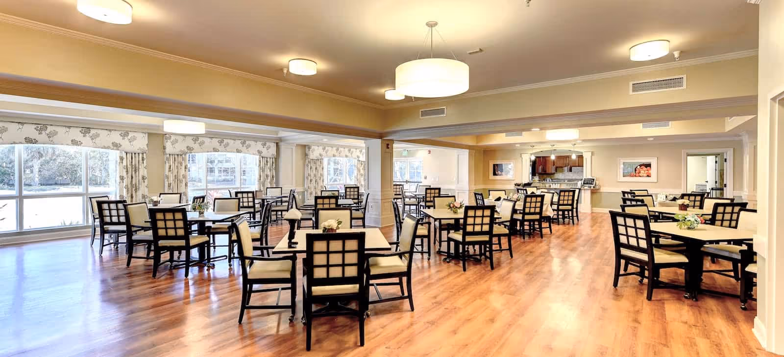 A spacious dining room with multiple square tables and cushioned chairs arranged neatly on a polished wooden floor. Large windows with floral curtains allow natural light to fill the room. Ceiling lights provide additional illumination, and small flower arrangements decorate each table. The room has a warm and inviting atmosphere.