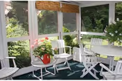 A bright sunroom with white wooden chairs and a round table surrounded by large windows showing green foliage outside. There are potted plants on the table and on the floor, and bamboo blinds partially cover the windows.