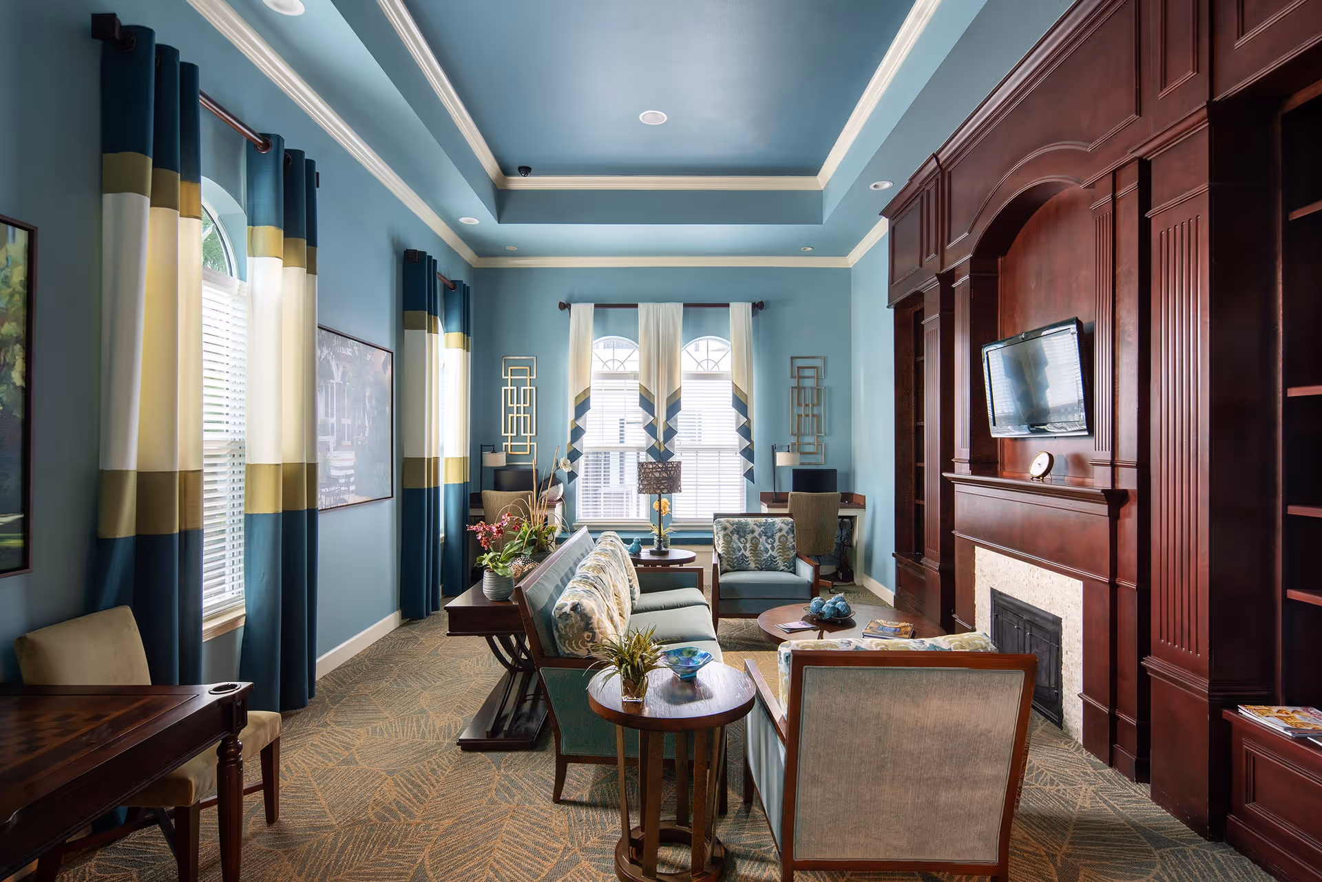 A cozy living room with blue walls and ceiling, featuring large windows with blue, white, and gold curtains. The room has a patterned carpet, several upholstered chairs and sofas with floral cushions, wooden side tables with plants and decorative items, and a large dark wood built-in fireplace with a mounted flat-screen TV above it.