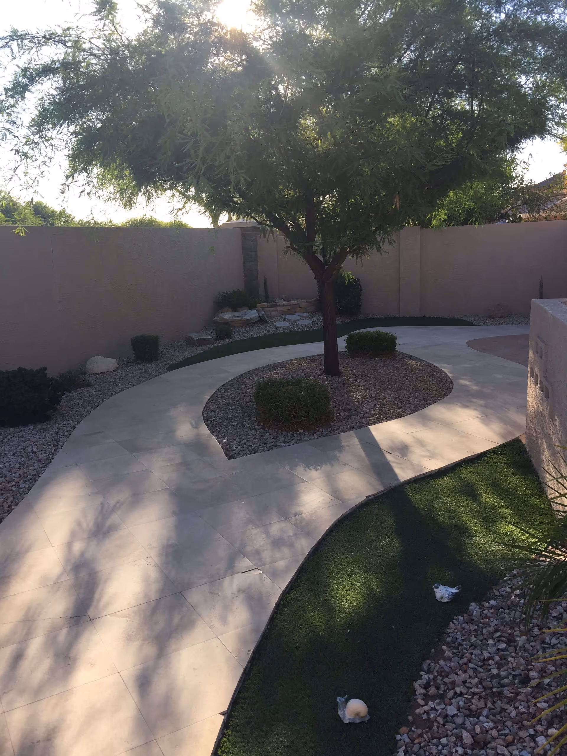 A curved paved walkway in an outdoor garden area with a tree in the center surrounded by small bushes and gravel. The area is enclosed by a beige wall, and there is artificial grass and decorative rocks along the path. Sunlight filters through the tree branches, casting shadows on the walkway.