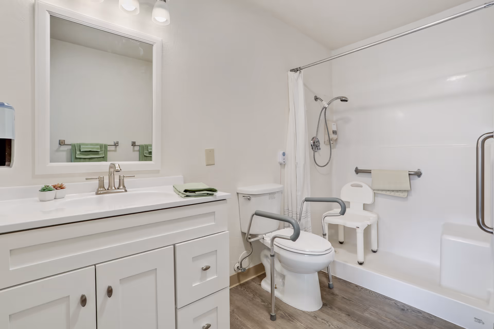 Bright accessible bathroom with a white vanity and mirror on the left, a toilet with grab bars in the center, and a tub/shower with a shower chair on the right.