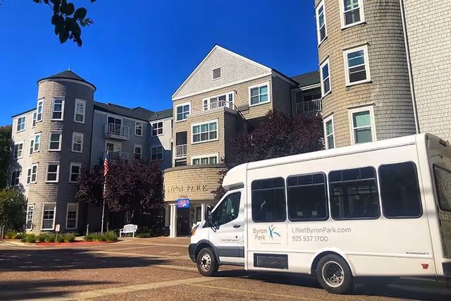 Exterior view of Byron Park senior living facility showing a multi-story building with gray siding and multiple windows. In front of the building, there is a white shuttle bus with Byron Park branding and contact information parked on a paved area. Trees and landscaping are visible around the building under a clear blue sky.
