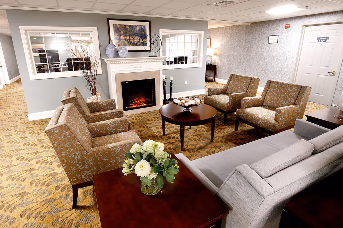 Comfortable senior living lounge with armchairs and a sofa arranged around a fireplace and central coffee table.
